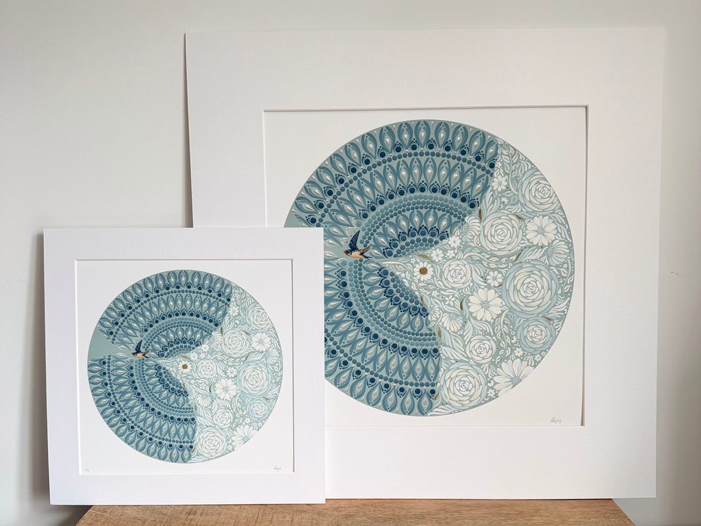 Two mounted  art prints with white mat boards, featuring a circular mandala-style illustration in shades of blue and white with small birds, displayed on a wooden surface against a light wall.